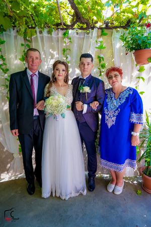 Formal wedding party gathering under leafy canopy with white backdrop.