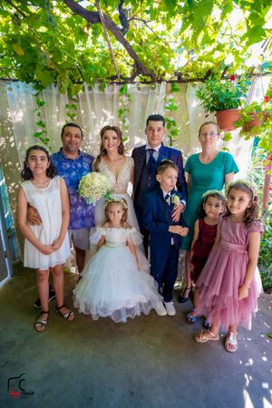 Family gathering under green vine-covered pergola wearing formal attire.