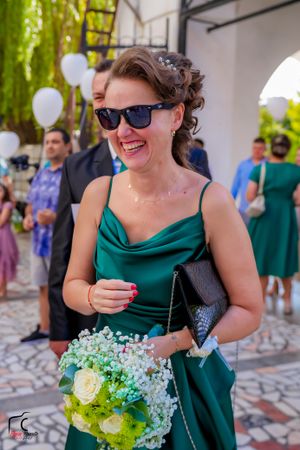 A woman in an emerald green dress holds a bouquet of white and green flowers at an outdoor event.