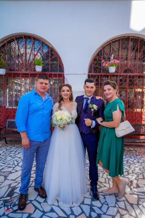 Wedding party members in formal attire pose against white church wall and red gates.