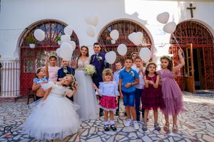 Wedding party and guests pose for photos against white church wall with red metal gates.