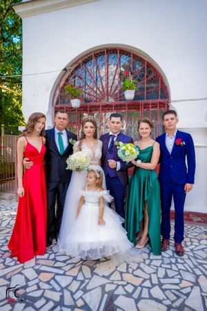 Wedding group photo showing attendees in formal attire of blue, green and red dresses against stone backdrop.
