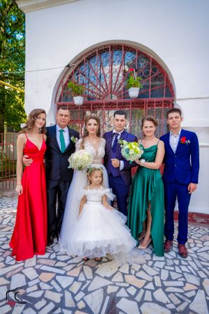 Family wedding portrait with bride in white gown and guests in colorful formal wear against arched backdrop.
