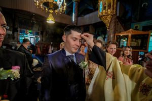 Orthodox Christian wedding ceremony shows priest blessing groom in navy suit during traditional religious service.