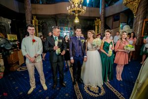 Wedding party members pose together on patterned blue carpet under elegant chandelier.