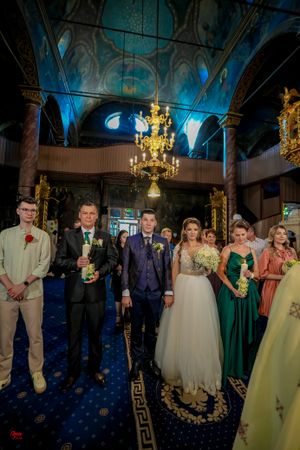 Wedding party stands in an ornate church interior with blue carpet and golden chandeliers overhead.