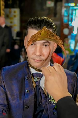 Groom wearing navy suit receives golden crown during Orthodox wedding ritual.