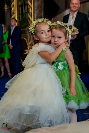 Two flower girls in white and green tulle dresses wearing floral crowns embrace at a wedding reception.