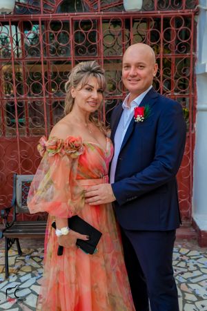 Couple in formal wear with coral dress poses against ornate iron gate background.