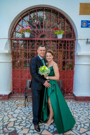 Formal couple in green dress and suit posing playfully by decorative gate.