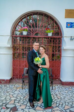 Couple in green formal wear and black suit pose with bouquet by stone archway.