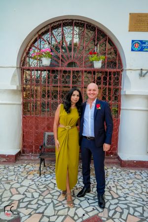 Couple in formal wear with mustard dress poses against white wall and red iron gate.
