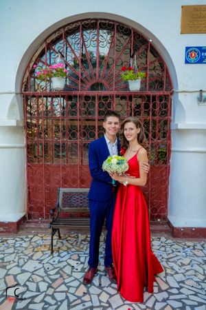 Couple in formal attire with red dress and blue suit pose by decorative gate.