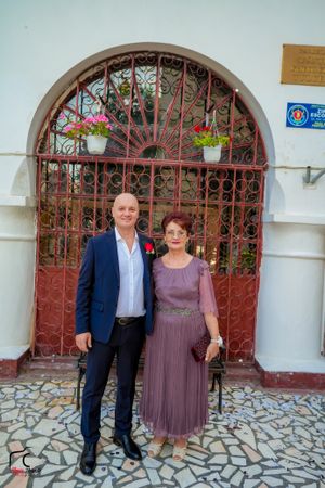 Couple poses in front of decorative red iron gates and stone floor wearing formal attire.