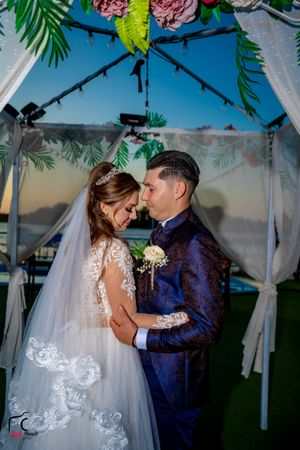 Wedding couple shares an intimate moment under a tropical-themed ceremony arch at sunset.