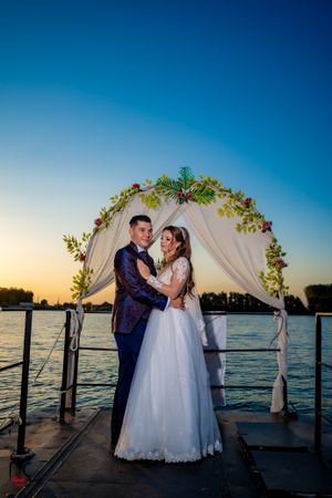 A romantic wedding ceremony on a waterfront dock at sunset under a floral arch with white draping.