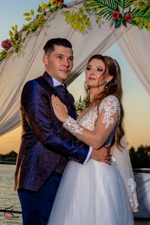 Elegant wedding portrait under floral arch with water backdrop at dusk.