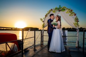 Wedding couple embracing under floral arch with dramatic sunset over water.