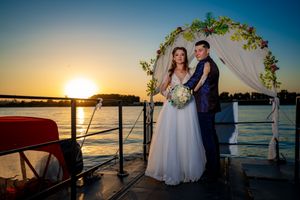 Wedding couple poses on boat deck with golden sunset and floral arch.