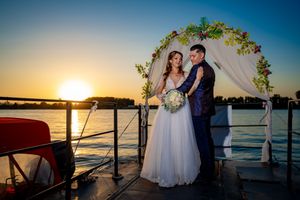 Wedding couple stands beneath decorated arch with sunset over water.