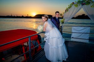 Wedding couple on decorated boat deck with white arch and vibrant sunset colors.