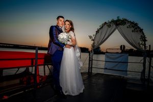 A couple poses in formal attire under a decorated arch at sunset by the water.