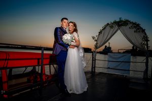 A formal couple embraces under a floral arch during sunset on a waterfront deck.
