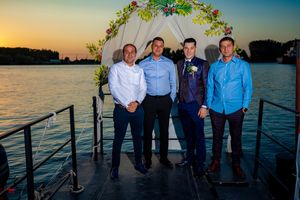 Four people in formal wear gather under a floral arch with water and sunset in background.