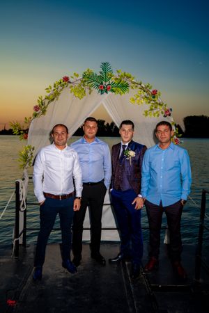 Four people in formal attire stand under a floral arch at sunset on a deck.