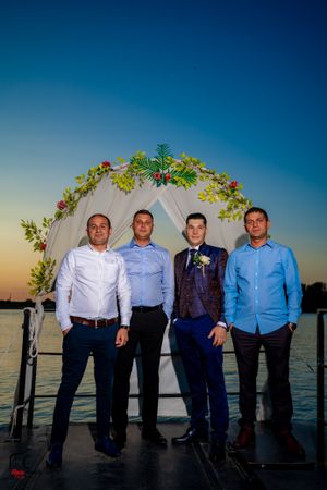 Four people in dress shirts pose under a decorated arch at dusk by the water.