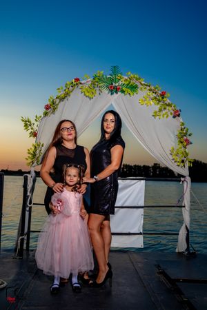 Three people pose together under a floral arch at sunset by the water.