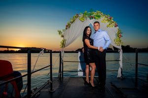 A couple poses against sunset waters under a floral wedding arch on a deck.