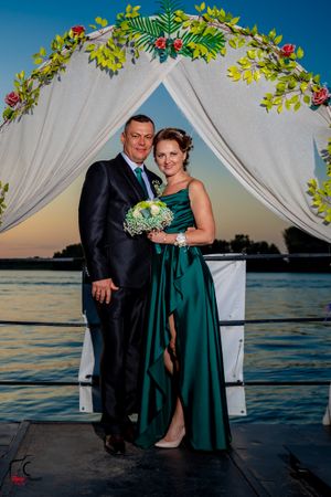 A couple poses under a floral arch at sunset on a waterfront deck for a formal event.