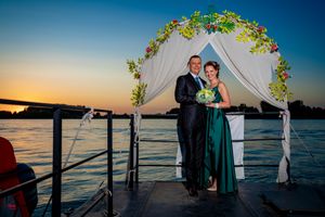 Wedding couple stands under decorated arch on waterfront dock during golden hour.