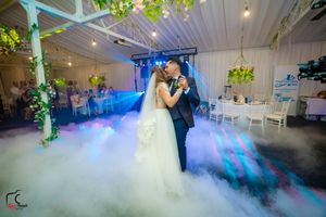 A wedding couple dances surrounded by atmospheric fog effects and colorful lighting in an elegant indoor venue.