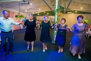 Line of dancers in formal attire holding hands under blue and purple lighting at an indoor venue.