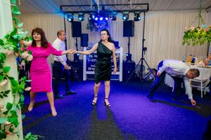 Dancers perform energetic moves on a blue-carpeted dance floor with stage lighting and decorative greenery.