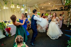 Wedding party dancing in a decorated reception venue with hanging lights and greenery.