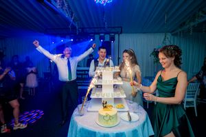 Wedding party gathers around tiered cake display during reception with vibrant blue lighting effects.