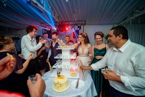 Wedding reception guests gather around an elegant white tiered cake in a dimly lit venue.