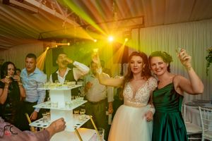 Wedding reception guests gather around a tiered white cake under warm yellow lighting at an indoor venue.
