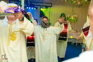 Musicians wearing cream-colored robes perform with drums during an indoor celebration.
