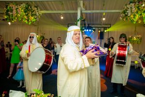 Performers in traditional Arab dress play drums and percussion instruments under decorative hanging foliage.