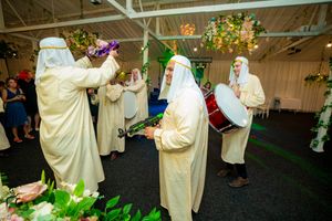 Musicians in flowing white robes perform with drums during a festive indoor event with decorative lighting.