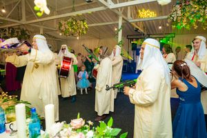 Performers in traditional Middle Eastern robes play drums and instruments at an indoor celebration with hanging greenery.