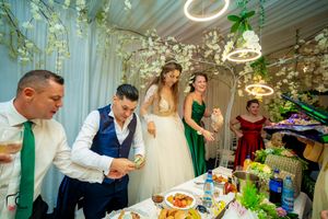 Wedding reception scene with guests dining at decorated table under hanging lights and greenery.