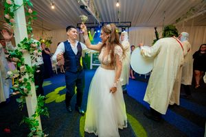Wedding reception guests dance and celebrate in a decorated indoor venue with green foliage and blue lighting.