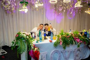 Elegant wedding reception head table decorated with floral arrangements and LOVE sign under purple lighting.