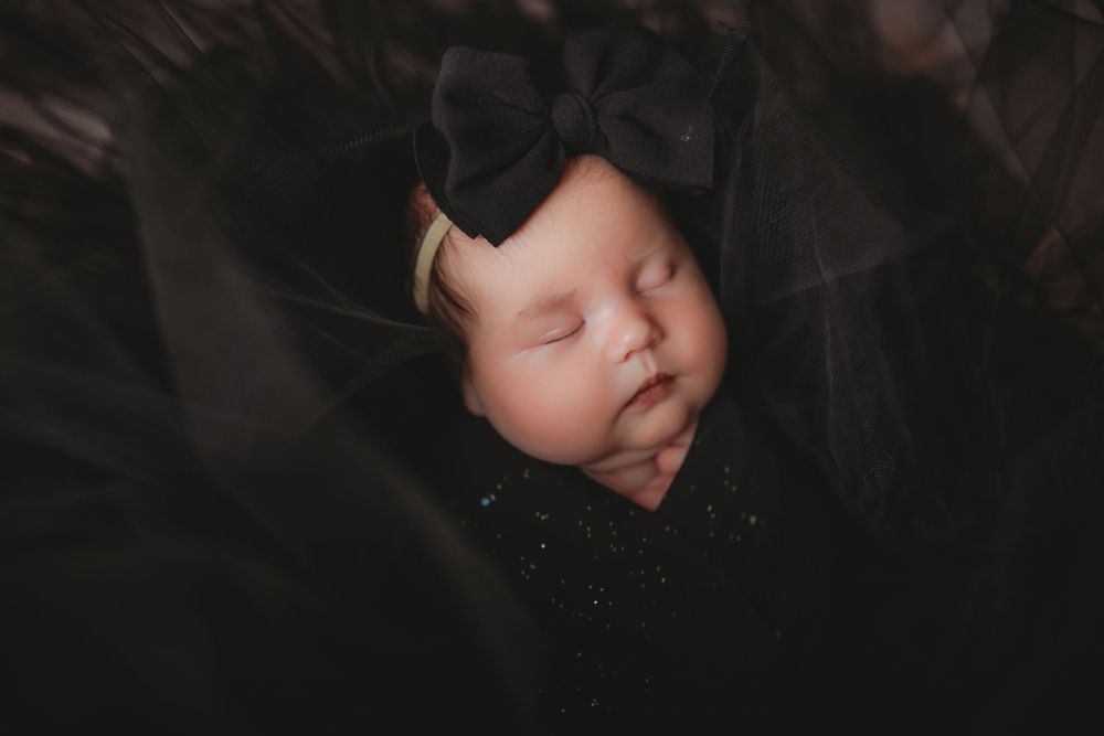 Sleeping newborn baby wrapped in black fabric with a black bow headband.