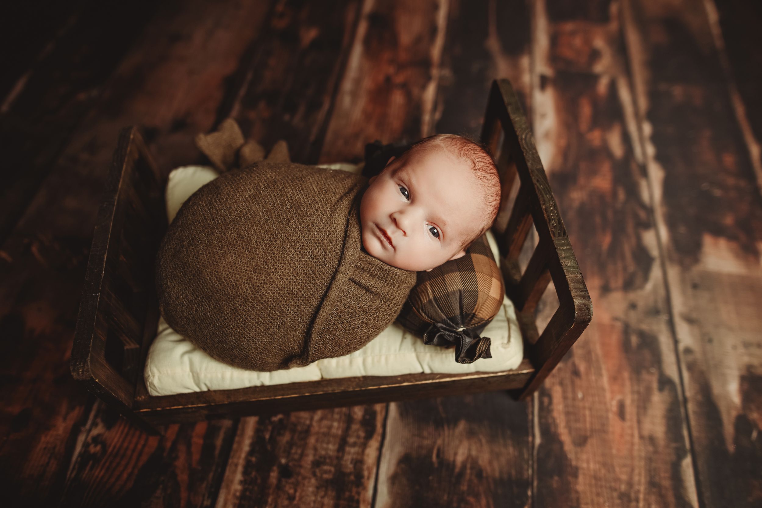 Newborn baby swaddled in brown wrap sleeping peacefully in a tiny wooden bed on rustic hardwood floors.